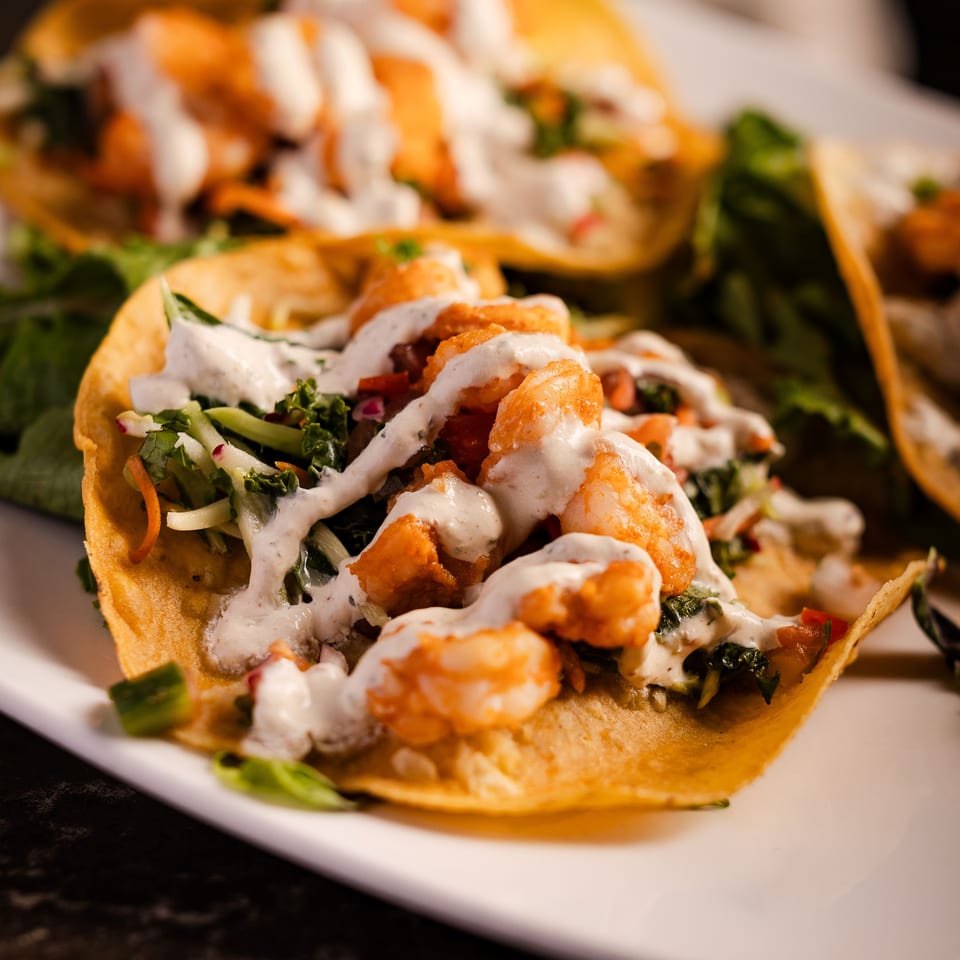 Shrimp tacos topped with creamy white sauce, chopped greens, and vegetables on a white plate.