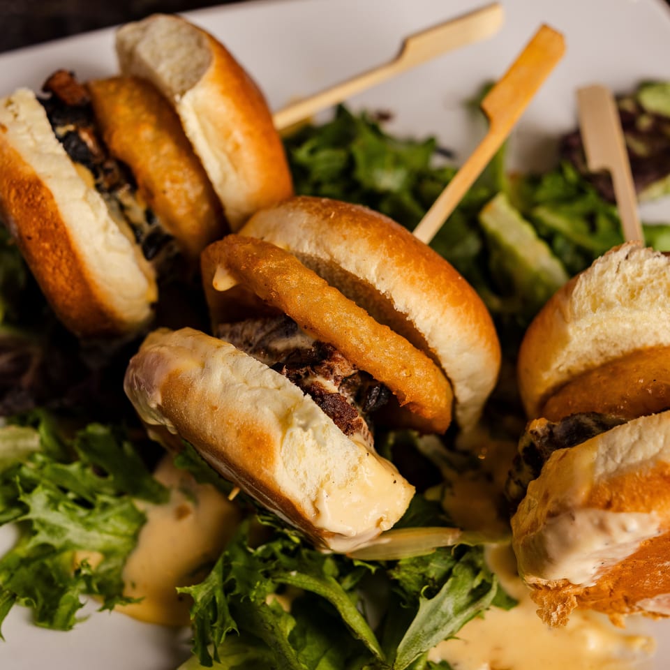 Mini burgers topped with fried onion rings arranged over leafy salad with creamy sauce on a white plate.
