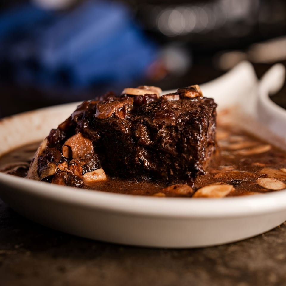Braised beef short rib topped with mushrooms and onions, served in rich sauce on a white plate.