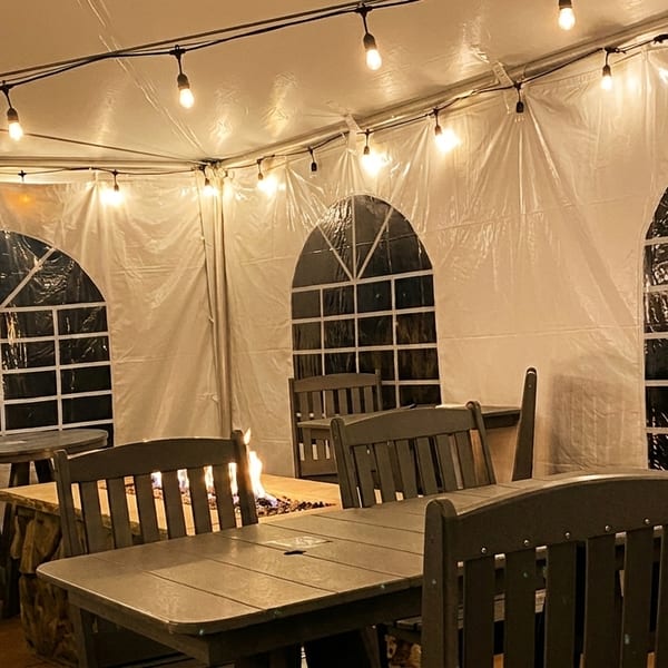 Outdoor dining tent with string lights, tables, chairs, and a fire pit, with a person seated.