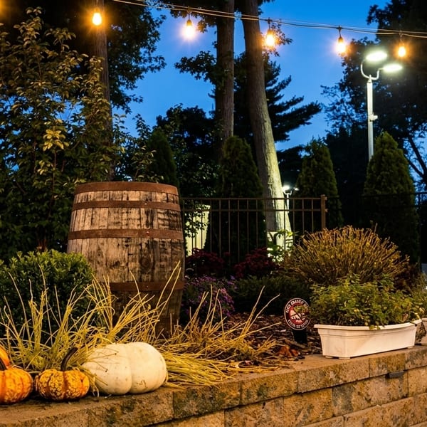 Outdoor patio at dusk with string lights, pumpkins, a barrel, and lush greenery.