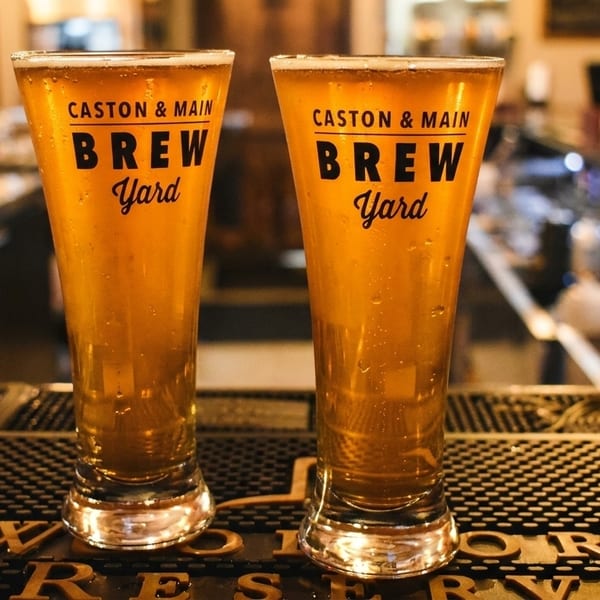 Two Caston & Main Brew Yard beer glasses filled with golden beer on a bar mat.