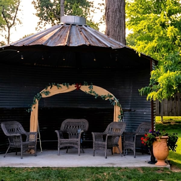 Outdoor seating area with wicker chairs under a rustic metal gazebo, surrounded by lush greenery.