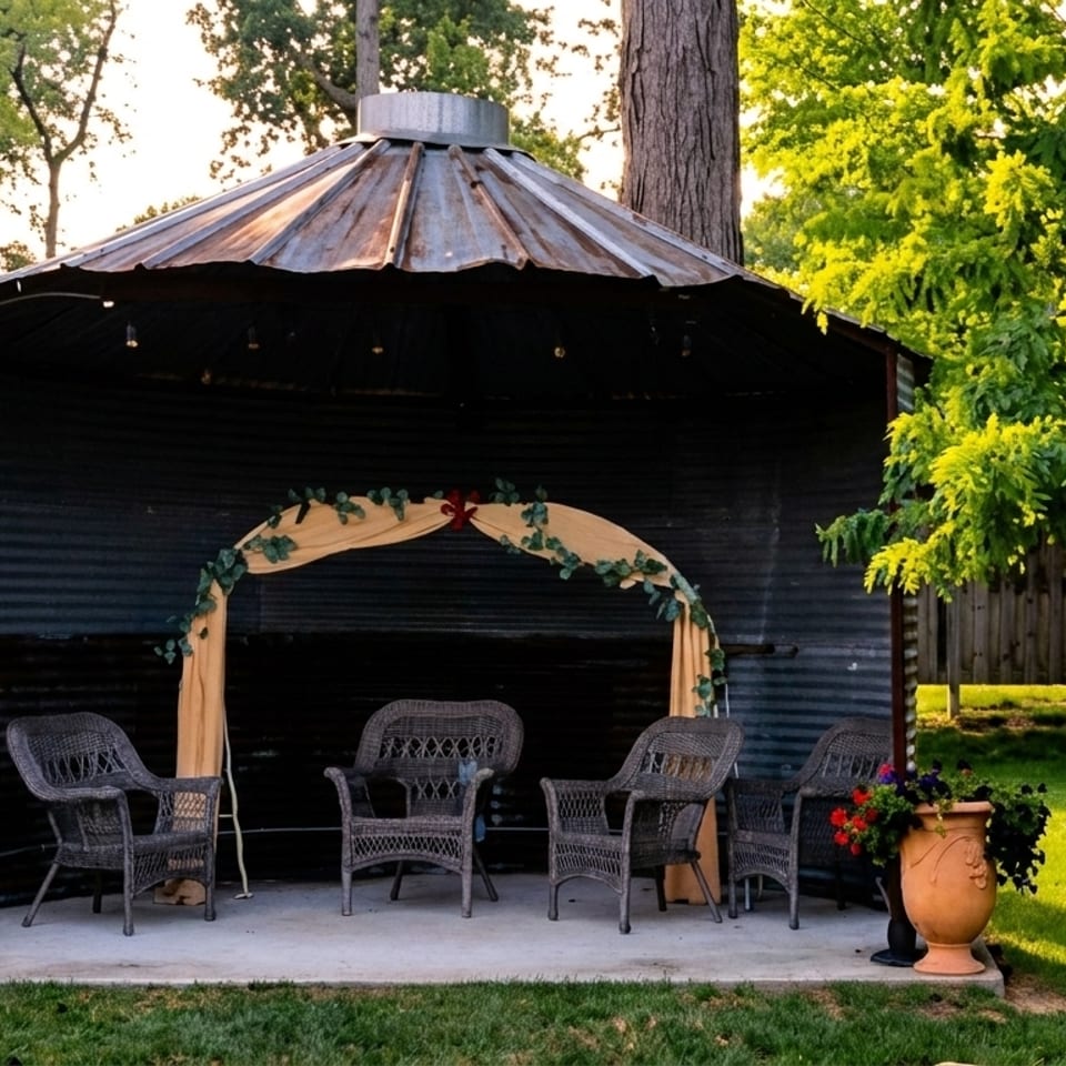 Outdoor seating area with wicker chairs under a rustic metal gazebo, surrounded by lush greenery.