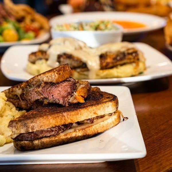 Grilled cheese sandwich with steak and mashed potatoes on a white plate, with other dishes blurred in the background.