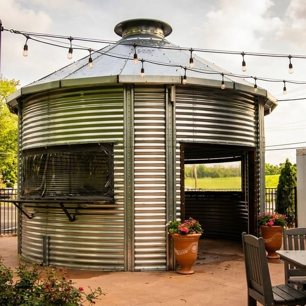 Outdoor patio with tables and chairs next to a corrugated metal silo structure under string lights.