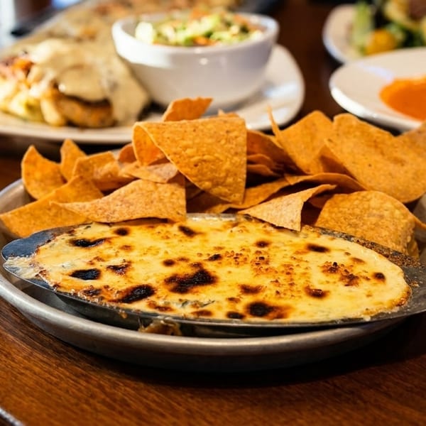 Cheesy baked dip with tortilla chips, alongside other dishes like soup and salad on a wooden table.