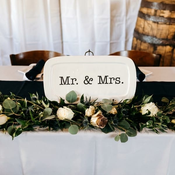 A "Mr. & Mrs." sign on a wedding head table with a floral garland and whiskey barrels.