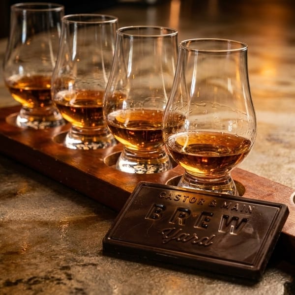 Four whiskey glasses on a wooden flight board with a "Brew Yard" chocolate bar on a stone counter.