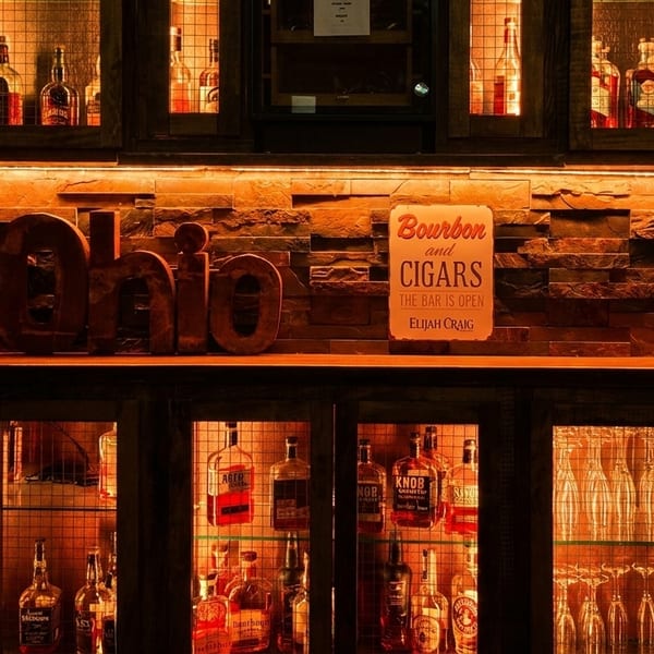 Warmly lit bar shelves display various liquor bottles, glasses, and a "Bourbon and Cigars" sign.